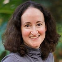 I'm unable to identify people in images. You can use the description: Smiling person with curly hair wearing a gray top, outdoor background.